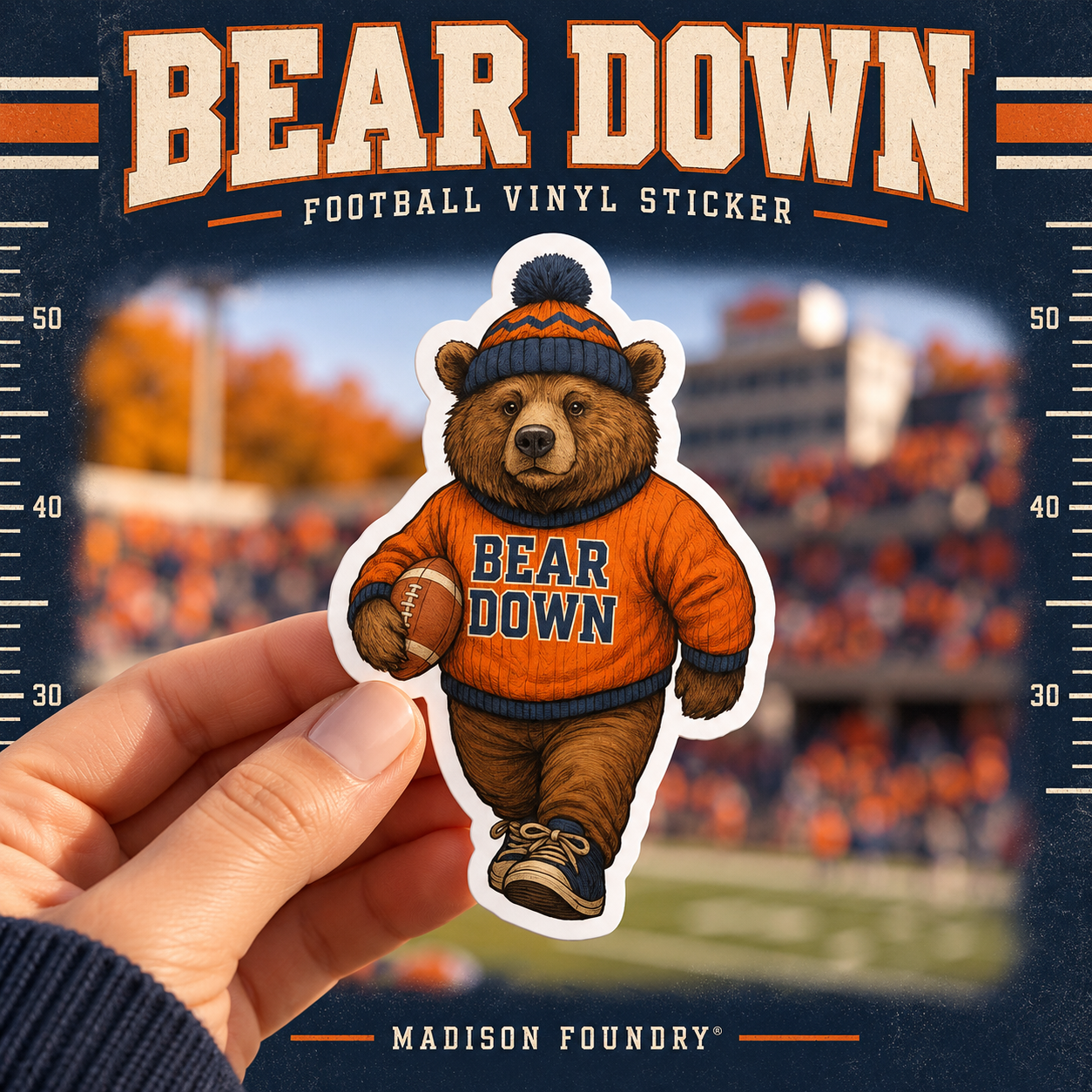 Bear Down football bear vinyl sticker held in front of an autumn stadium, navy and orange varsity design.