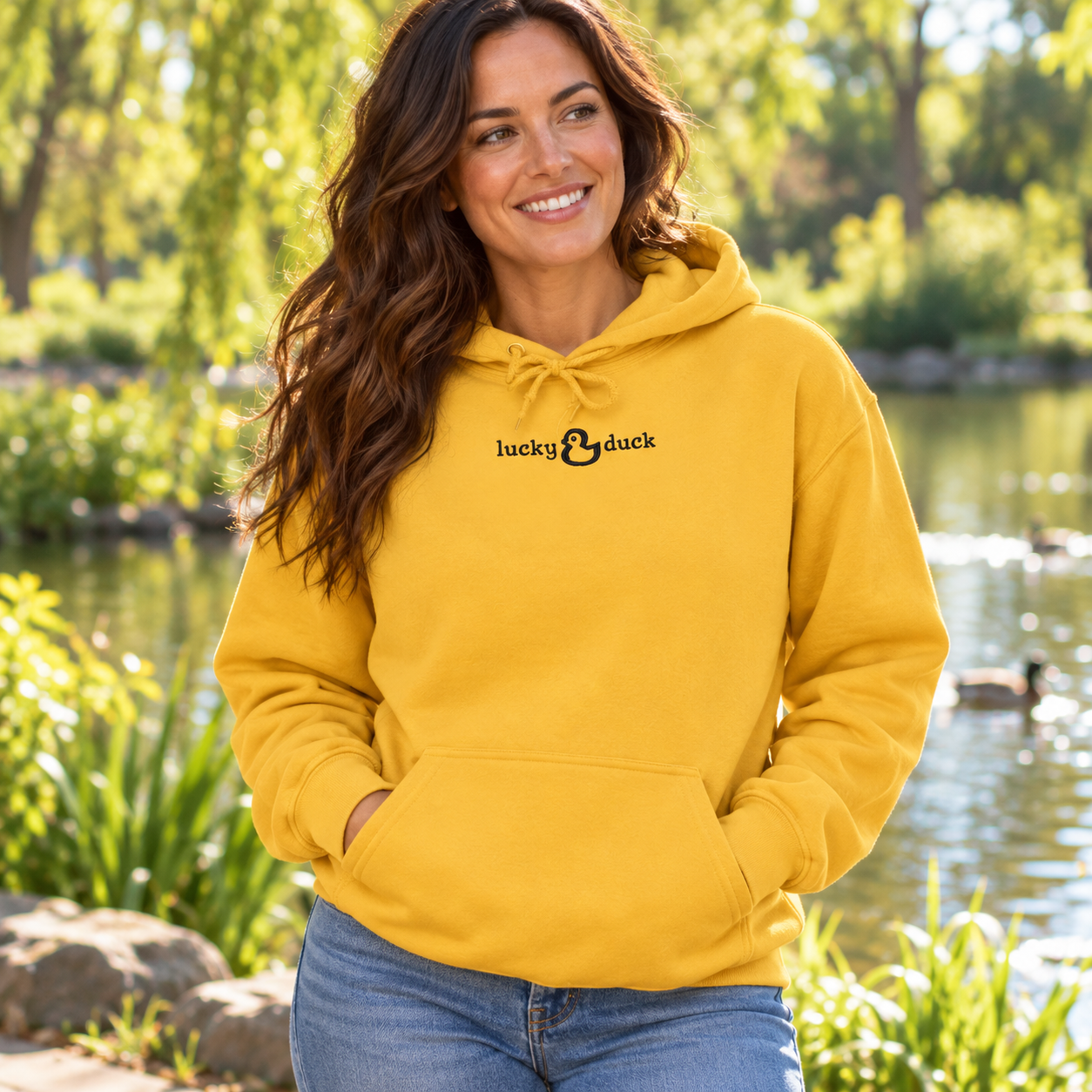 Woman wearing Gold Lucky Duck embroidered hoodie beside a sunny park pond